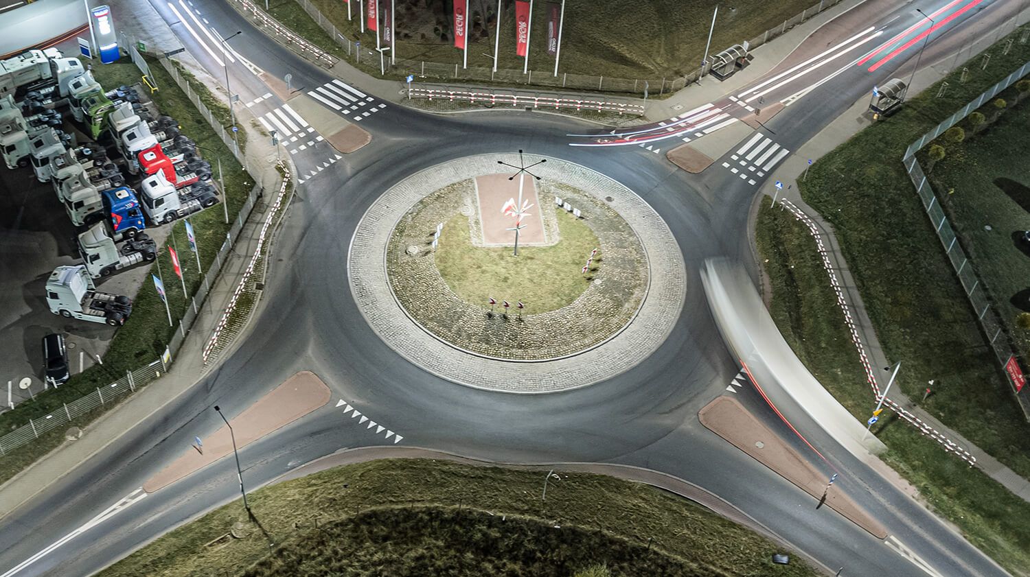 Noční pohled z ptačí perspektivy na kruhový objezd osvětlený pouličními lampami, zachycující automobilový provoz a dopravní značení vyznačující směr jízdy.